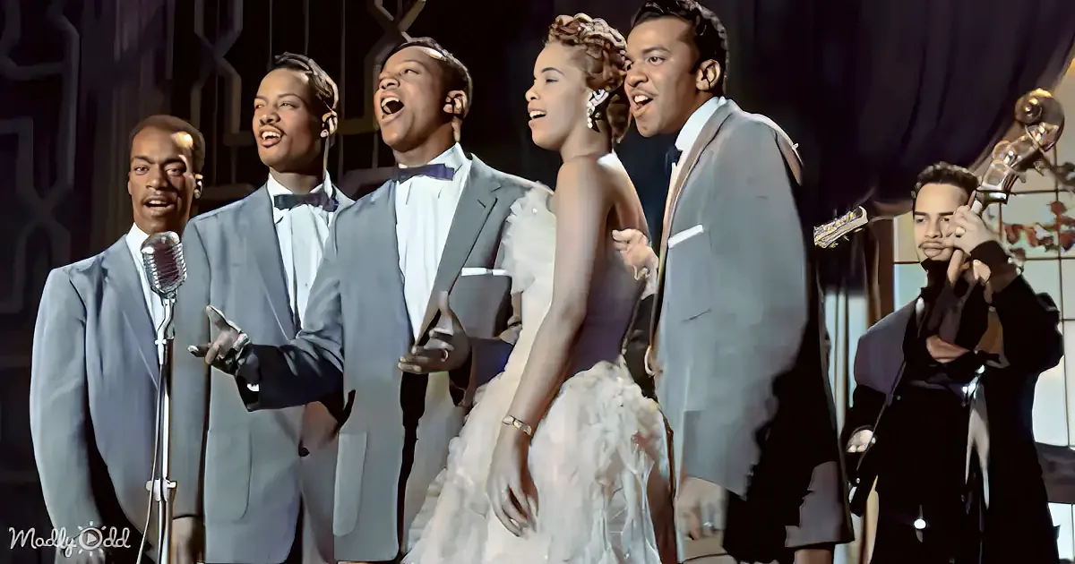 A group of five men in suits sing, accompanied by a woman in a white dress and a musician playing a bass.