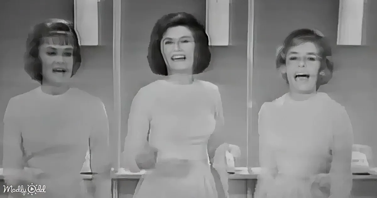 Three women in white dresses stand in a kitchen, smiling and appearing to sing or speak.