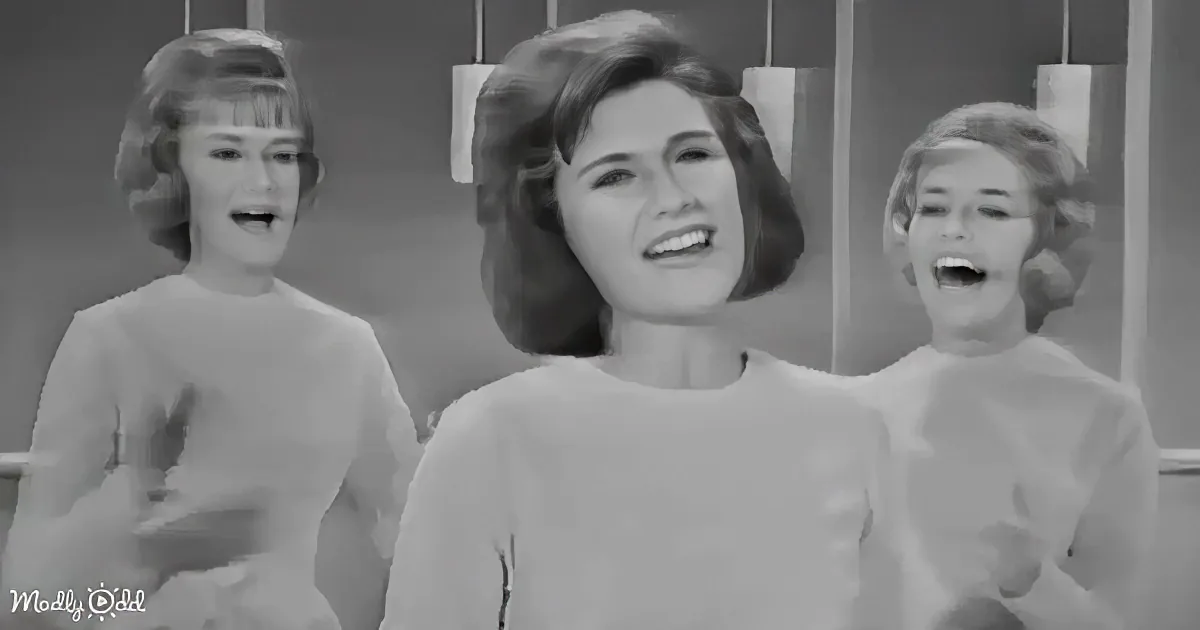 Three women in white shirts sing together, their expressions animated, in a vintage black-and-white setting.