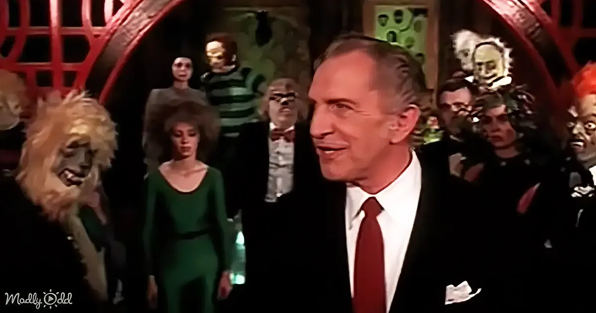A man in a suit and red tie stands in front of a group of people in costumes and masks.