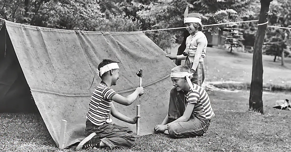 Children in striped shirts and headbands play with sticks near a tent in a grassy area.
