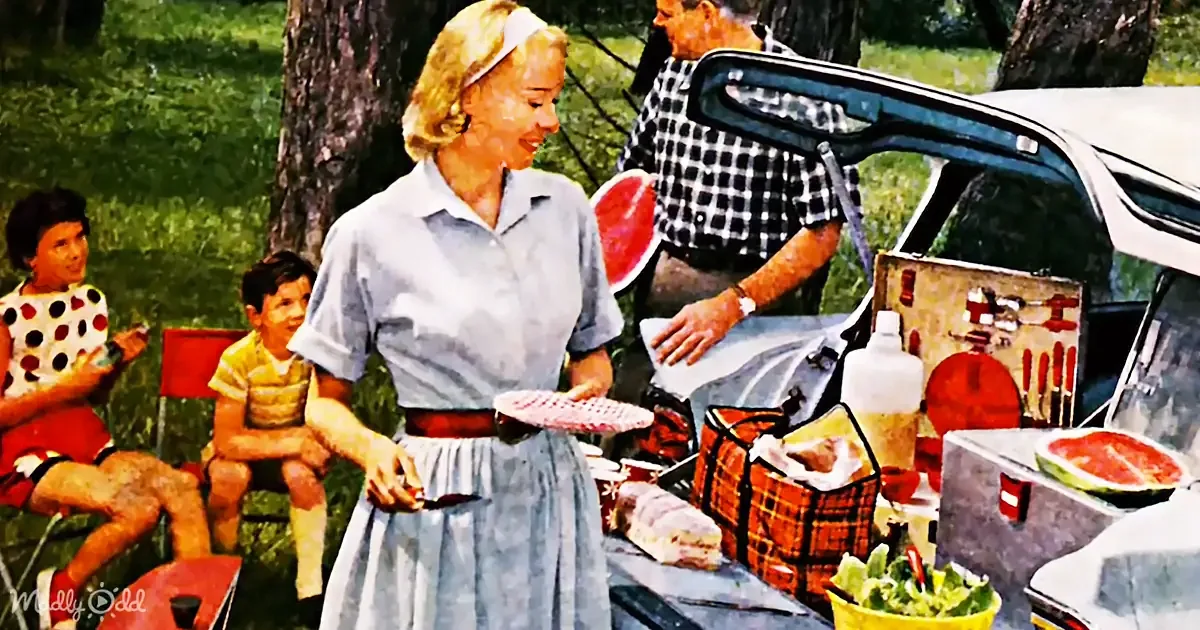 A woman in a blue dress holds a red plate, preparing food at a picnic with a man and children in the background.