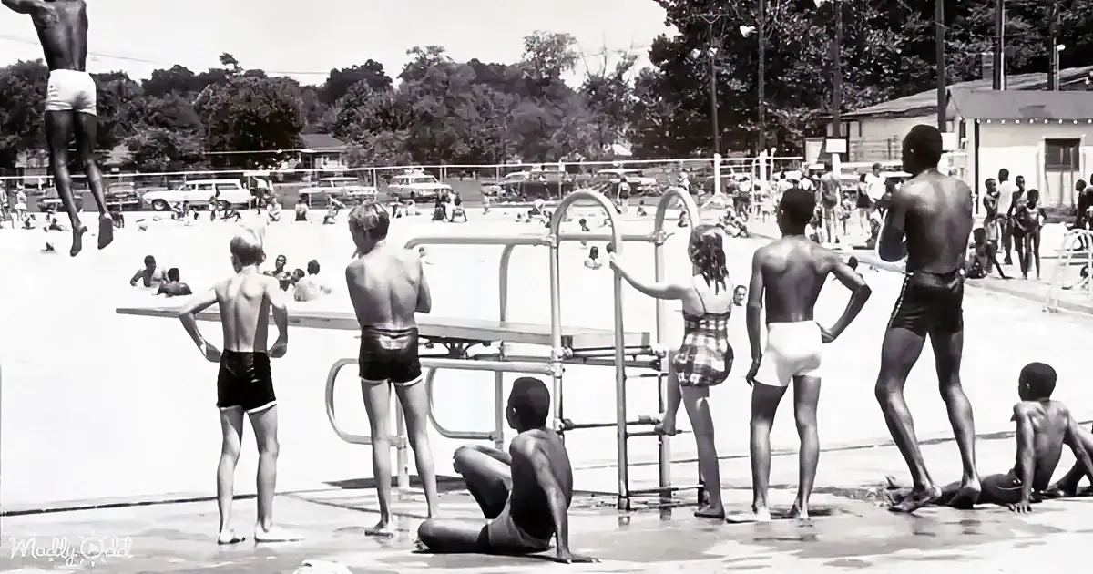 A boy jumps into a crowded swimming pool, surrounded by other swimmers and onlookers.