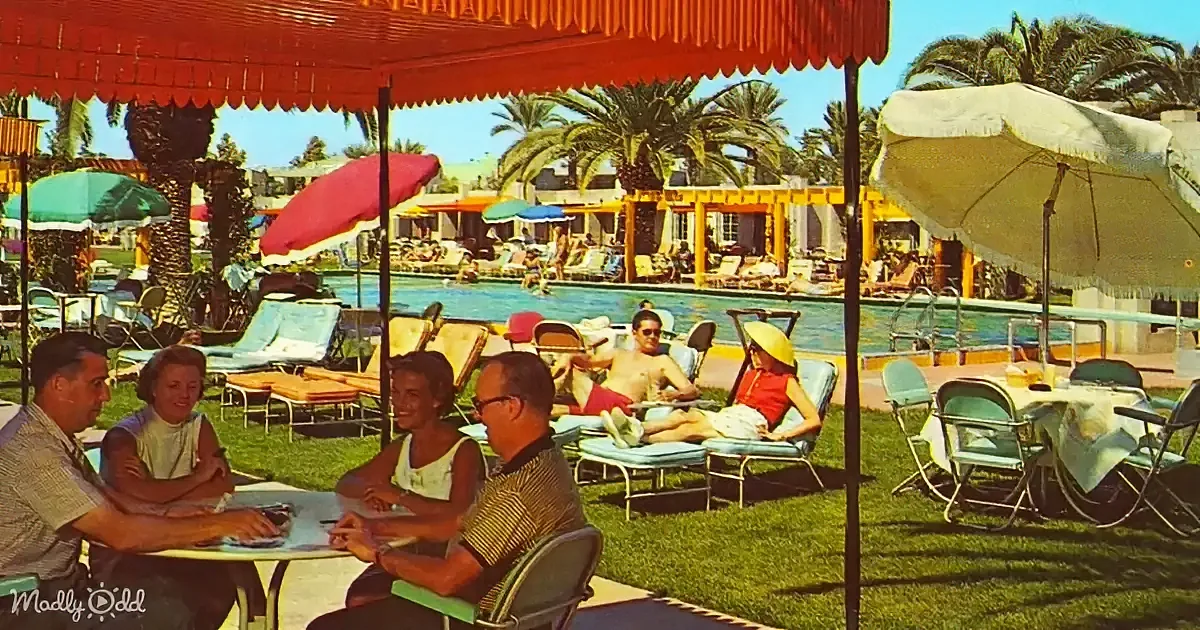 People relaxing by a poolside with colorful umbrellas and palm trees in the background.