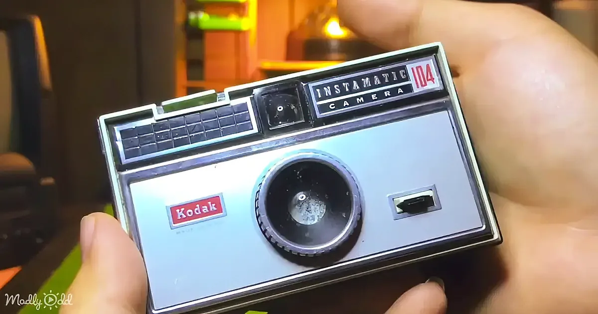 A woman with red nails holds a vintage Instamatic camera, smiling with a blue background.