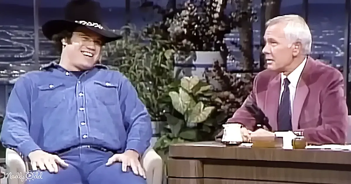John Wayne in a cowboy hat laughs while seated next to a man in a suit on a talk show set.