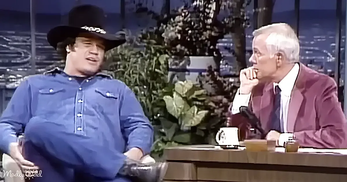 A man in a cowboy hat and blue shirt sits on a couch, talking to a man in a suit on a talk show set.