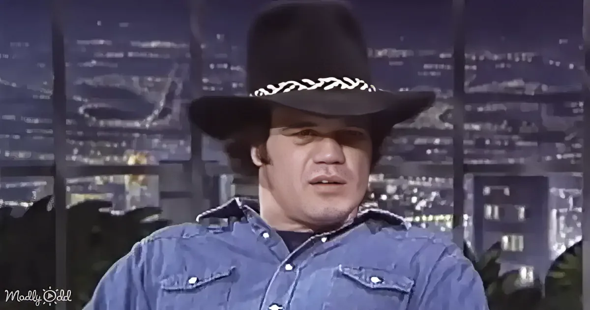 A man in a denim shirt and black cowboy hat stands against a cityscape backdrop.