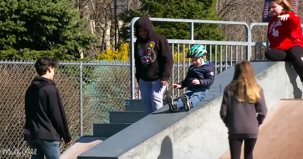 A child in a helmet sits on a concrete ledge at a skate park, surrounded by onlookers and trees.
