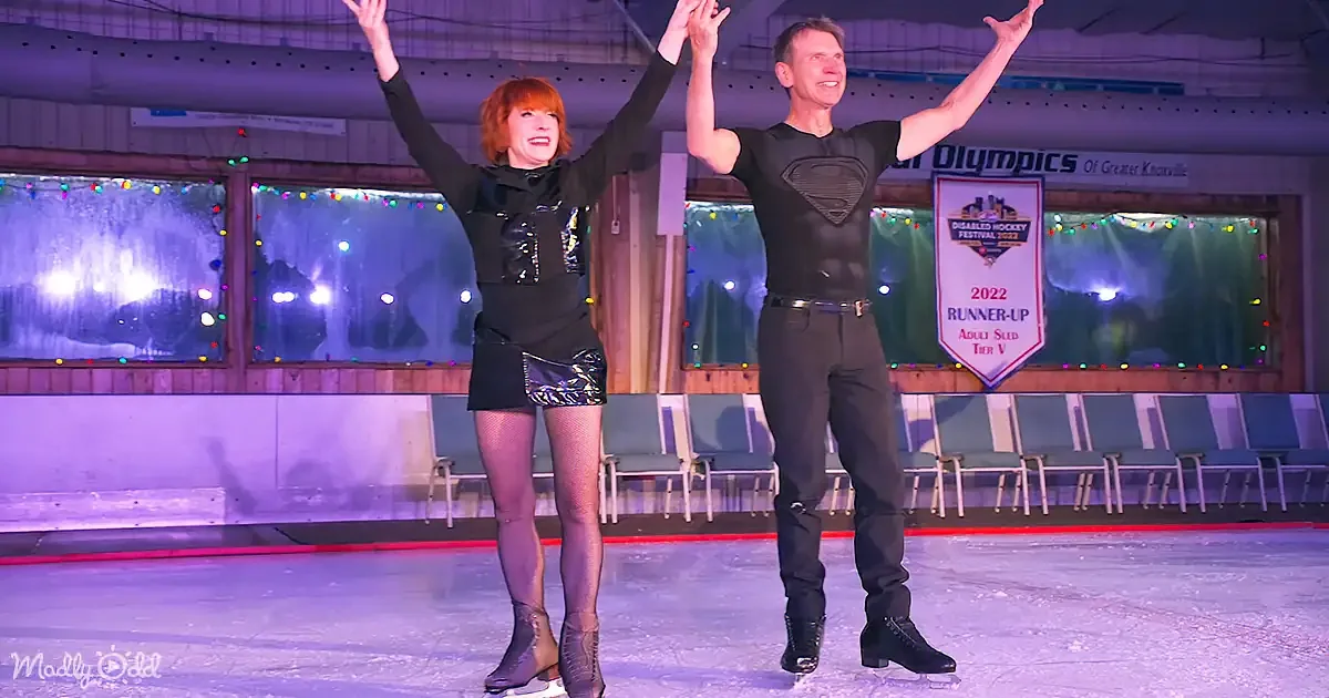 A man and woman in black ice skate on an ice rink, arms raised, celebrating.