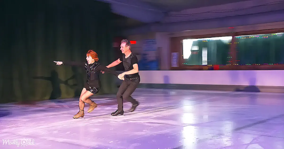 A man and woman skate together on an ice rink, holding hands, with the woman pointing forward.