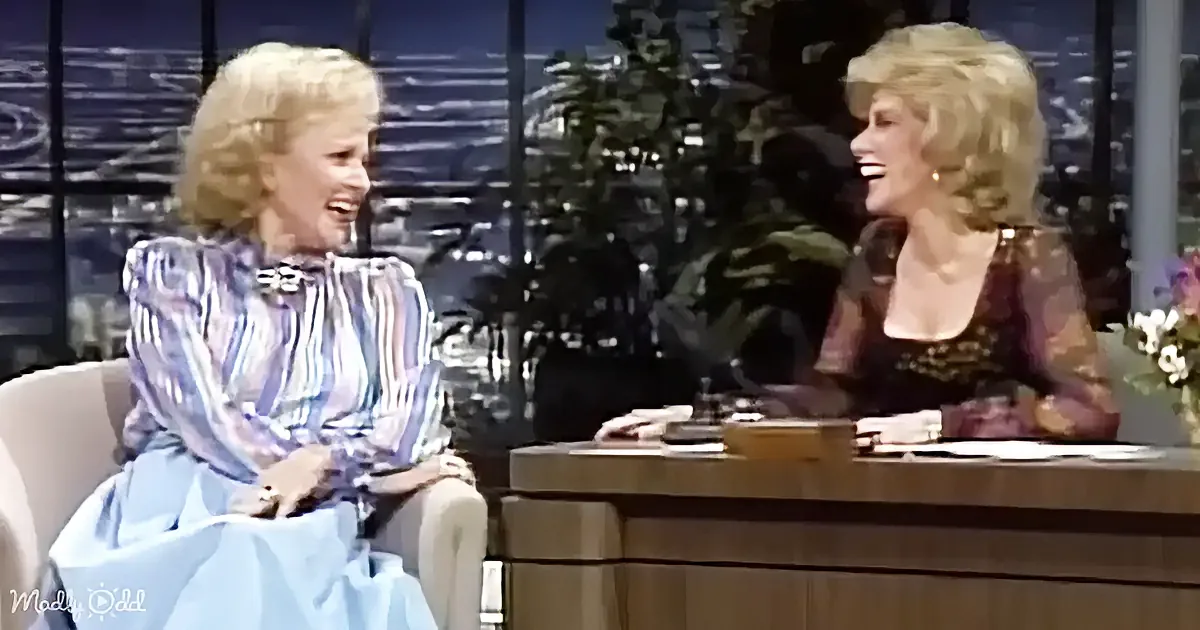 Two women are laughing on a talk show set, one in a blue dress, the other in a patterned top.