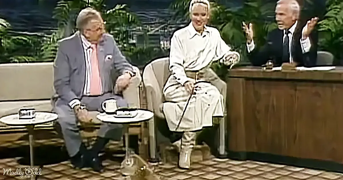 A man in a suit sits on a couch, gesturing with his hands, while a woman in a white outfit sits beside him.