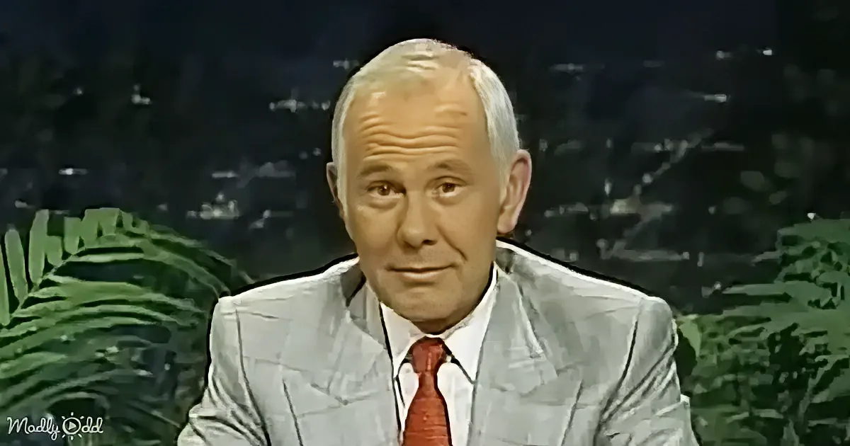 A man in a light gray suit and red tie sits in front of a dark background with green foliage.