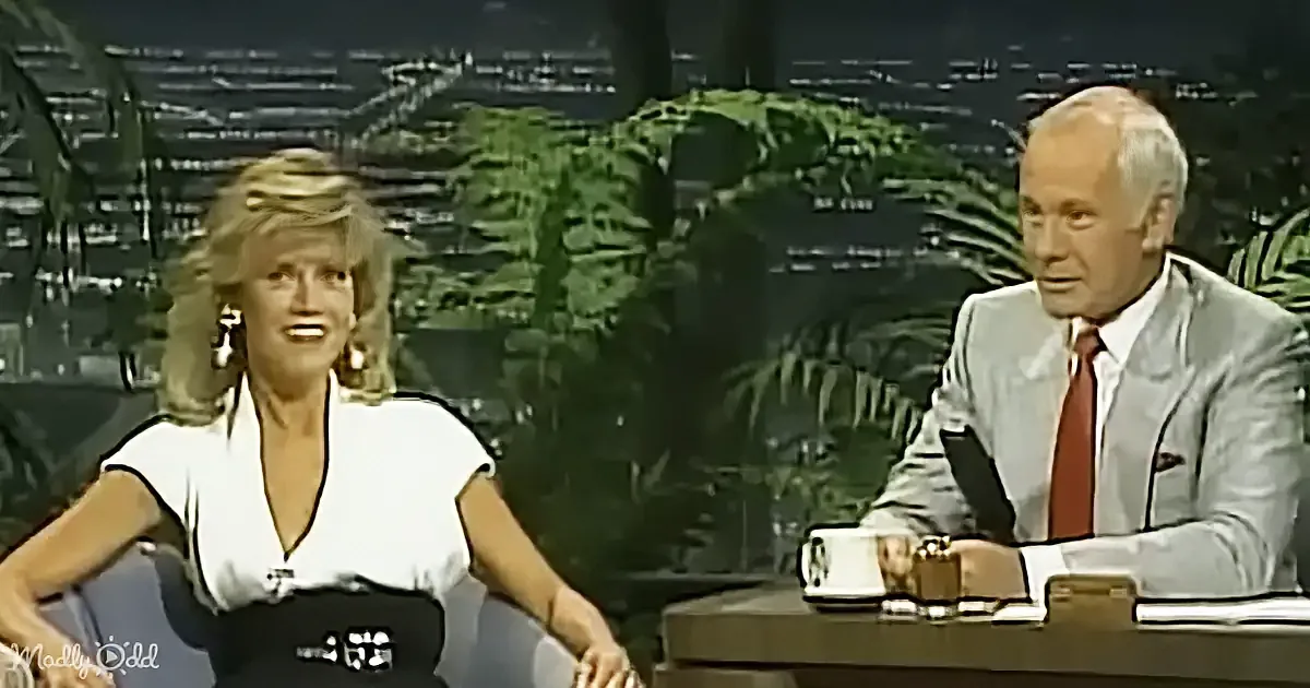 A woman in a white dress smiles while seated next to a man in a suit on a talk show set.