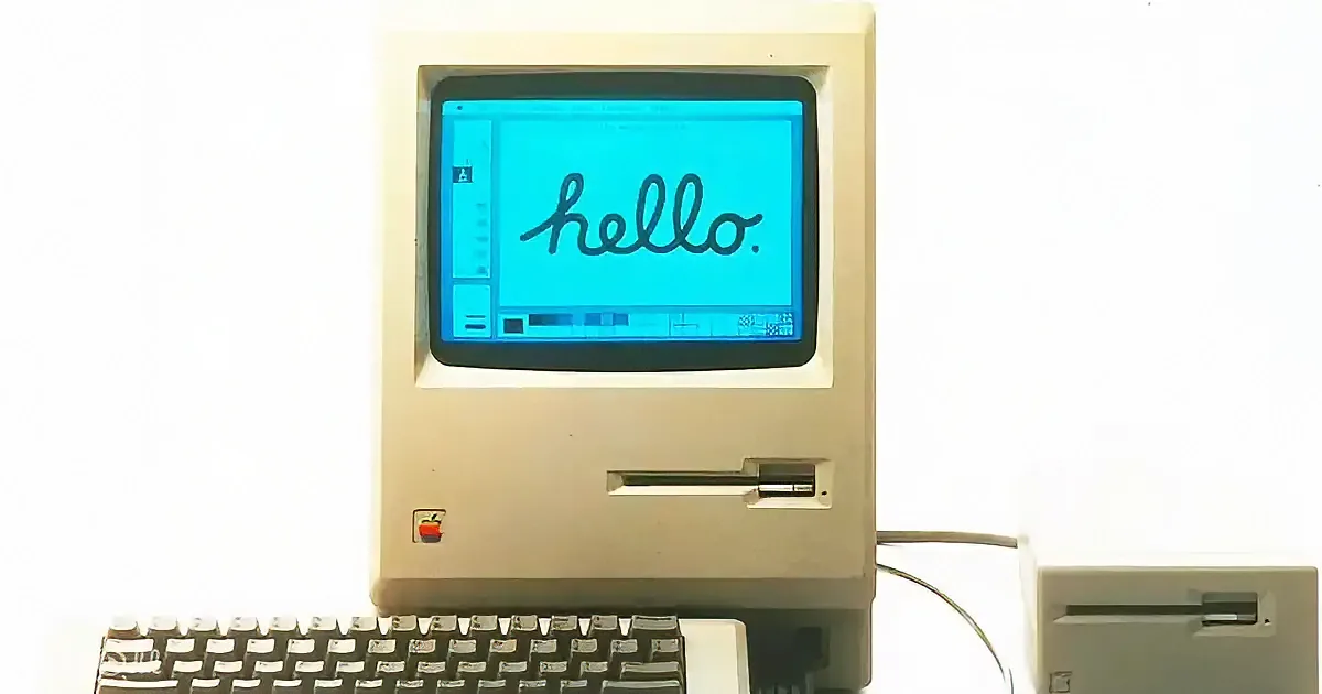 An old Apple computer displays "hello." on its screen, accompanied by a keyboard and a floppy disk drive.