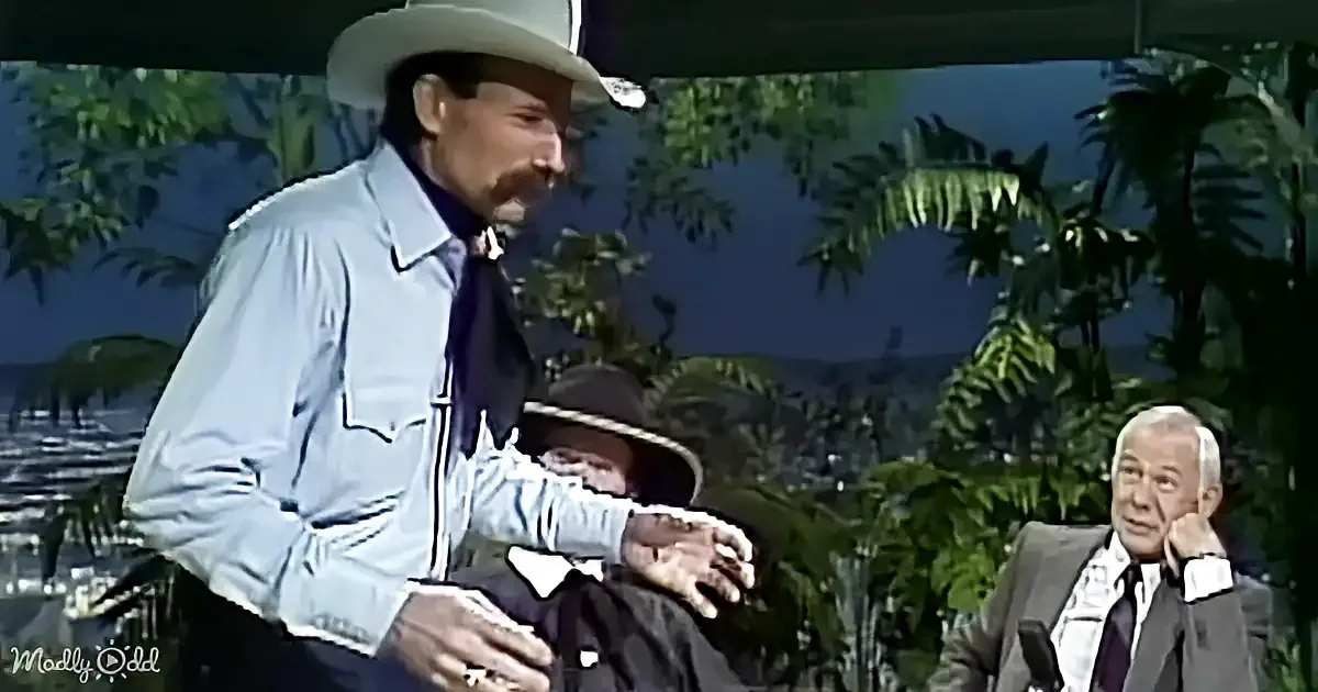 A man in a cowboy hat and light blue shirt gestures with his hands while standing outdoors.