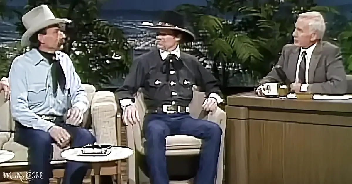 Three men in cowboy attire sit on a talk show set, one in a suit, discussing.