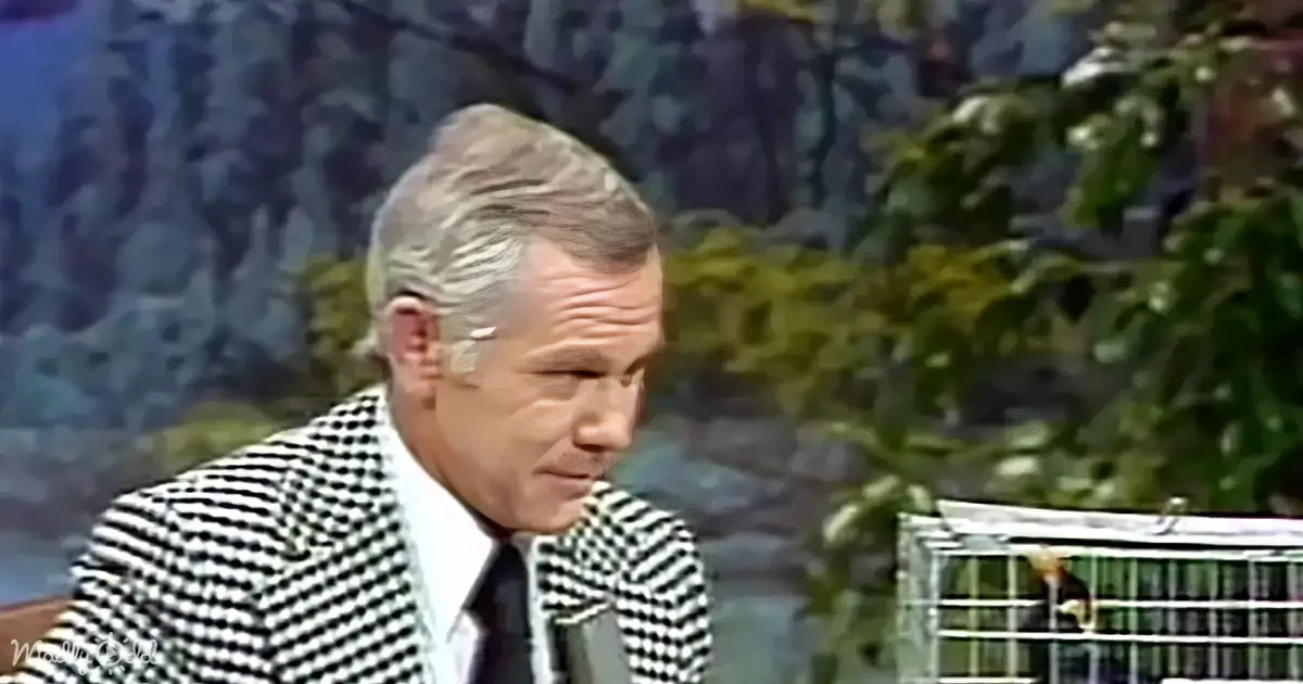 A man in a checkered suit jacket and tie is seated, looking to the side, with a birdcage and greenery in the background.