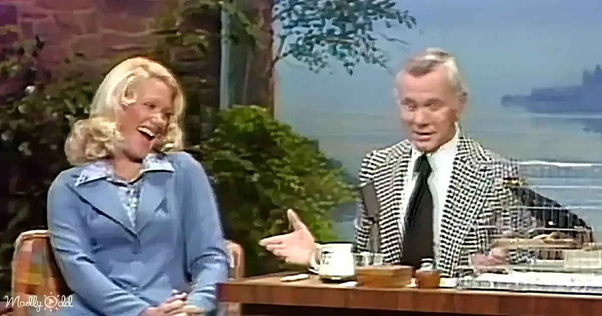 A woman in a blue dress laughs while seated next to a man in a checkered suit on a talk show set.