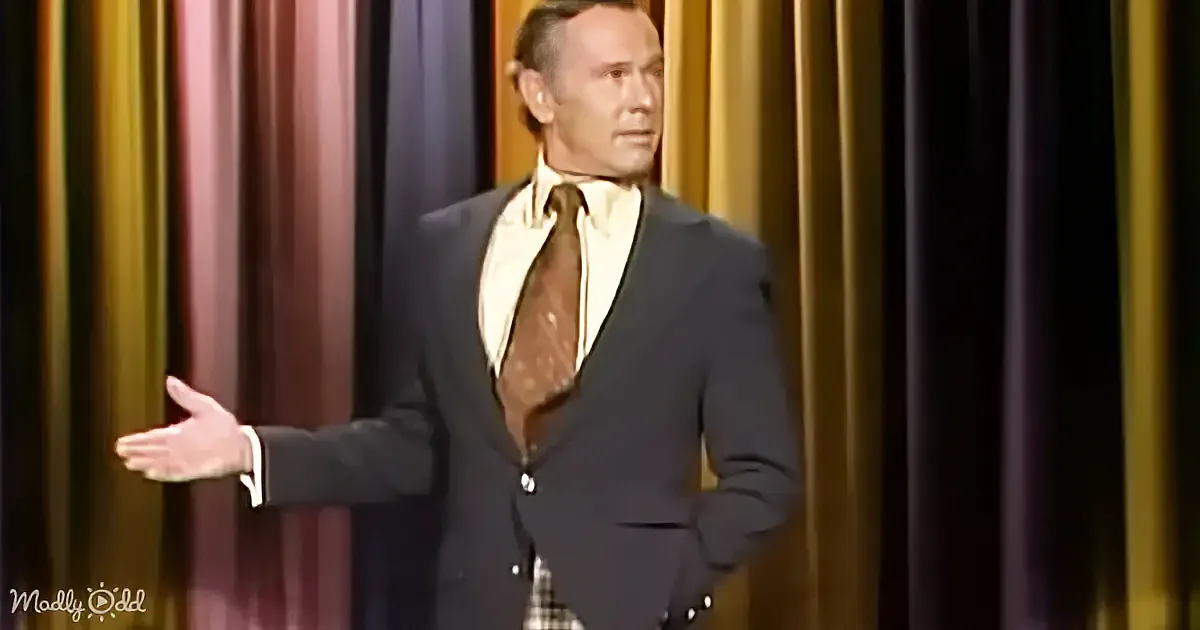 A man in a suit and tie gestures with his right hand, standing in front of a colorful curtain backdrop.