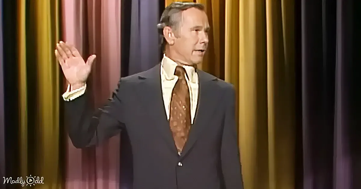 A man in a suit and tie raises his hand, standing against a colorful curtain backdrop.