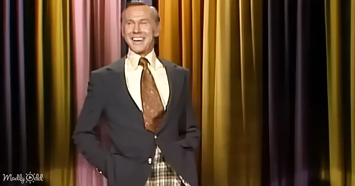 A man in a suit and tie smiles on stage with a colorful curtain backdrop.