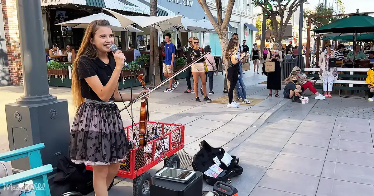 A girl in a black dress sings into a microphone while holding a violin, surrounded by people on a busy street.