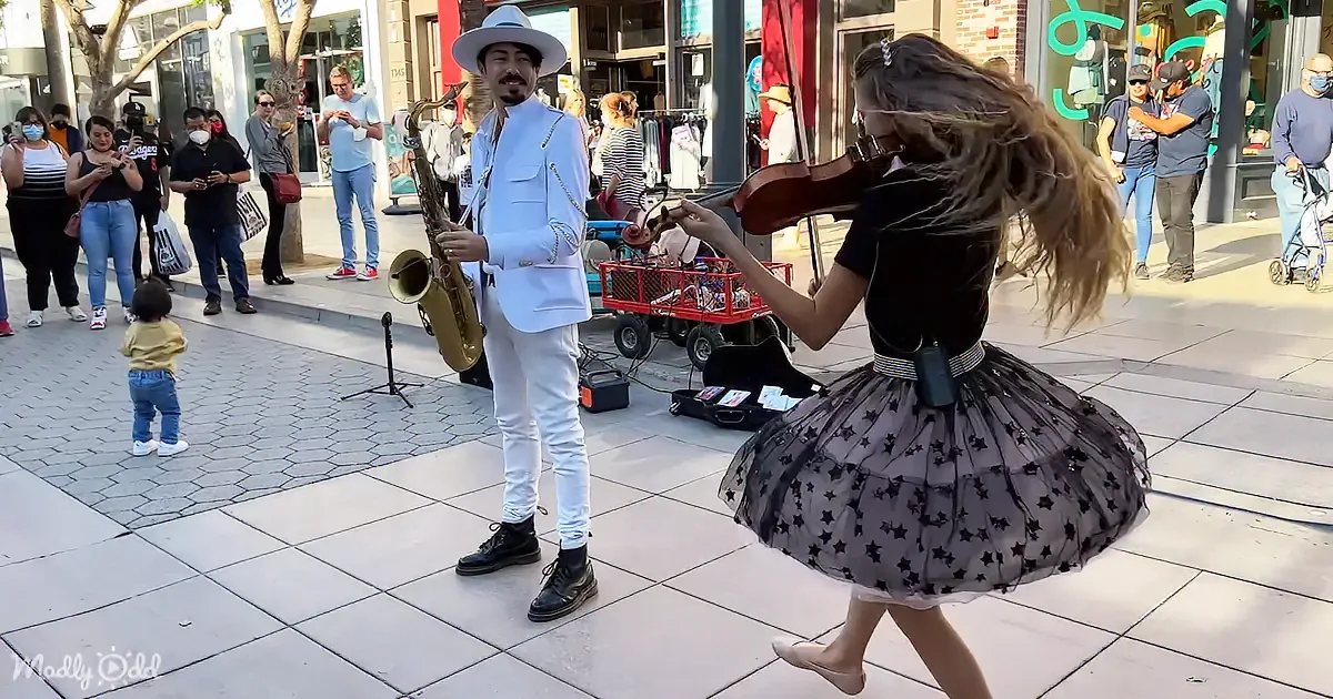 A man in a white suit plays saxophone while a woman in a black tutu dances nearby on a city sidewalk.