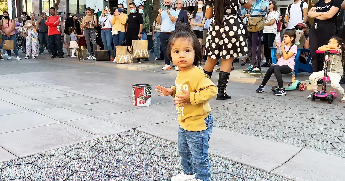 A toddler in a yellow hoodie stands on a paved area, surrounded by a crowd of onlookers.
