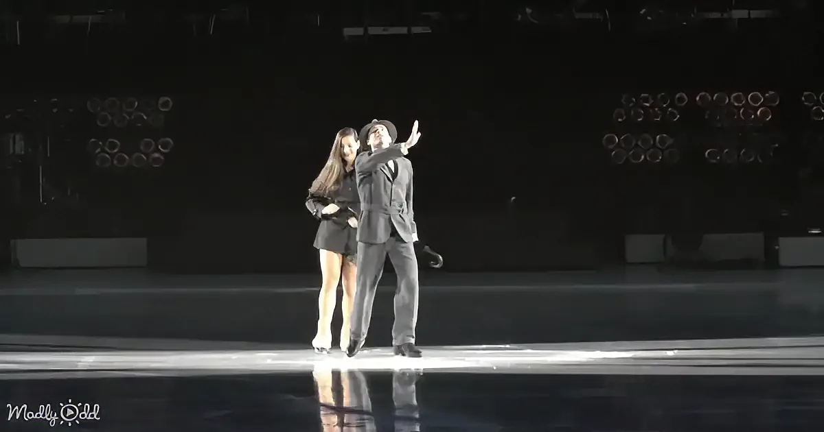 A couple in formal attire dances gracefully on a stage with spotlights in the background.