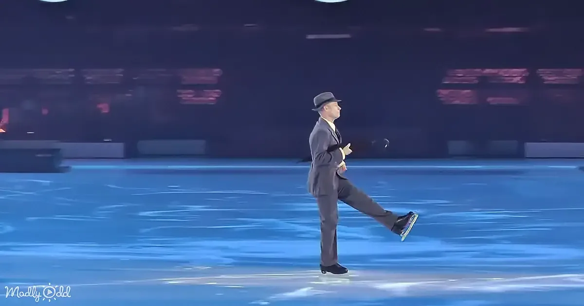 A man in a suit and hat skates on an ice rink, performing a leg kick.