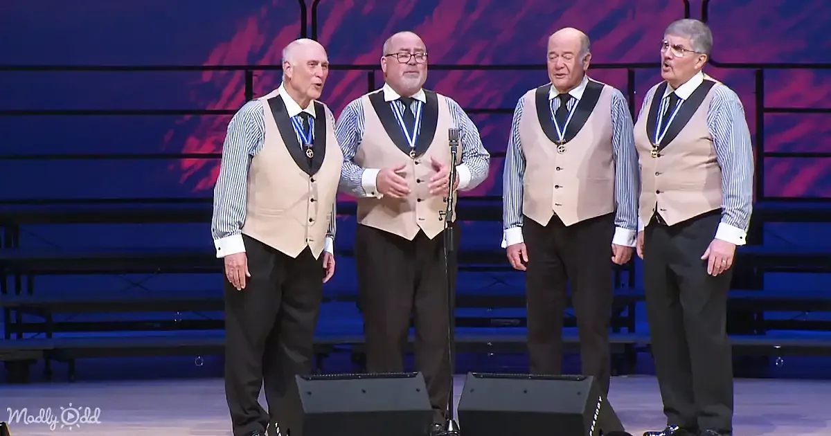 Four men in beige vests sing on stage, wearing medals and ties, with a microphone in front of them.