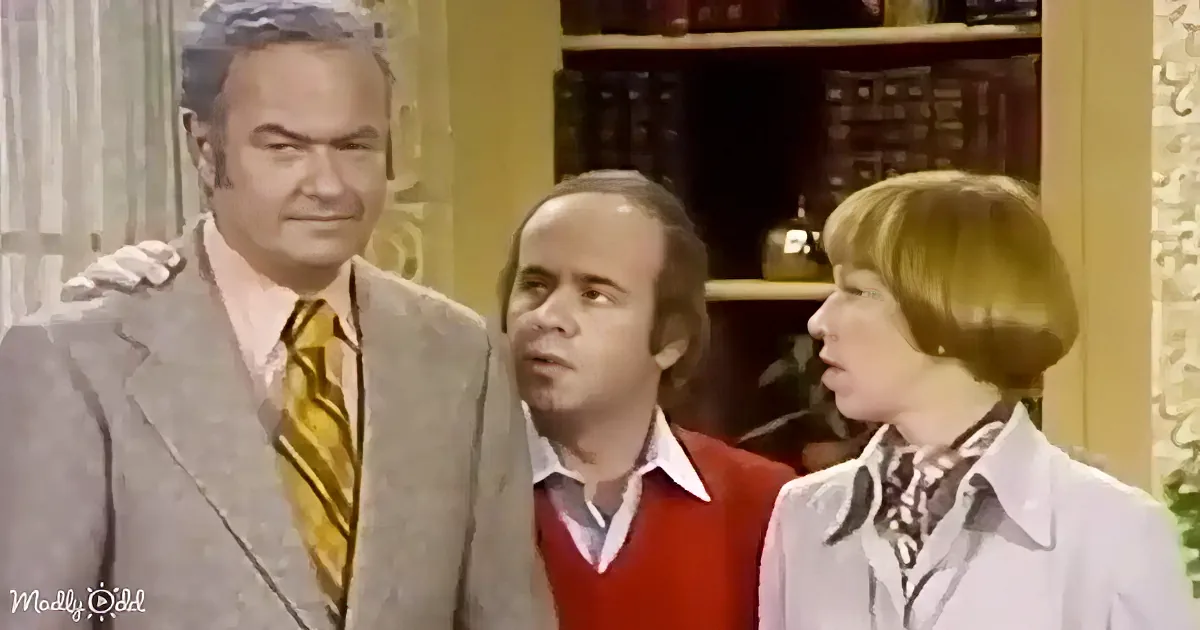 A man in a suit and tie stands with another in a red vest and a third in a white coat, all in a room with bookshelves.