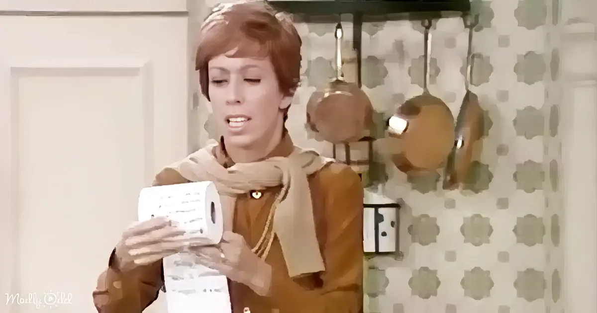 A woman in a brown jacket and scarf holds a roll of paper towels in a kitchen with patterned wallpaper.