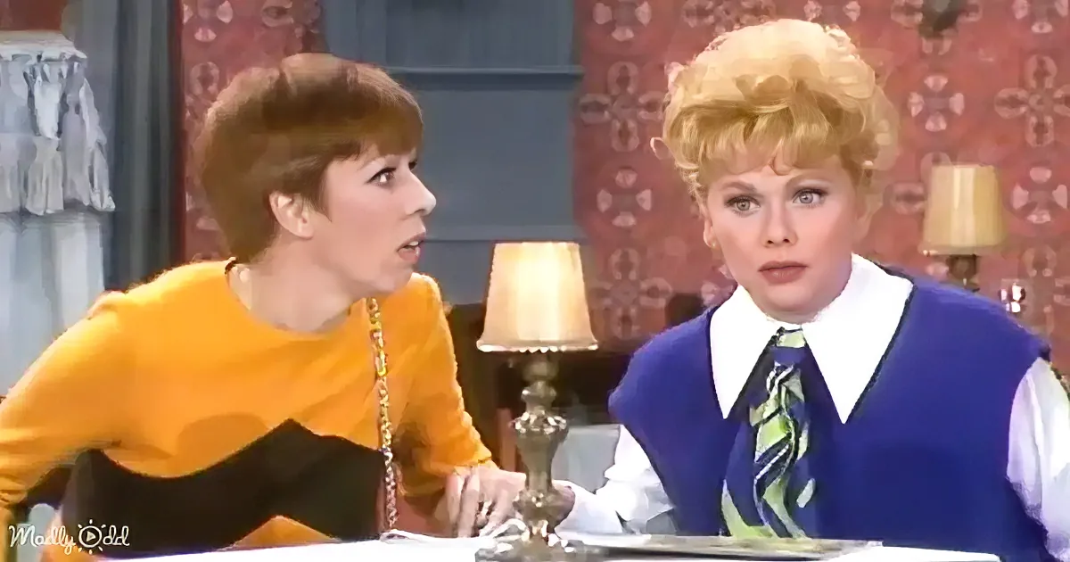 Two women in vintage attire are seated at a table, engaged in conversation, with a lamp and patterned wallpaper in the...