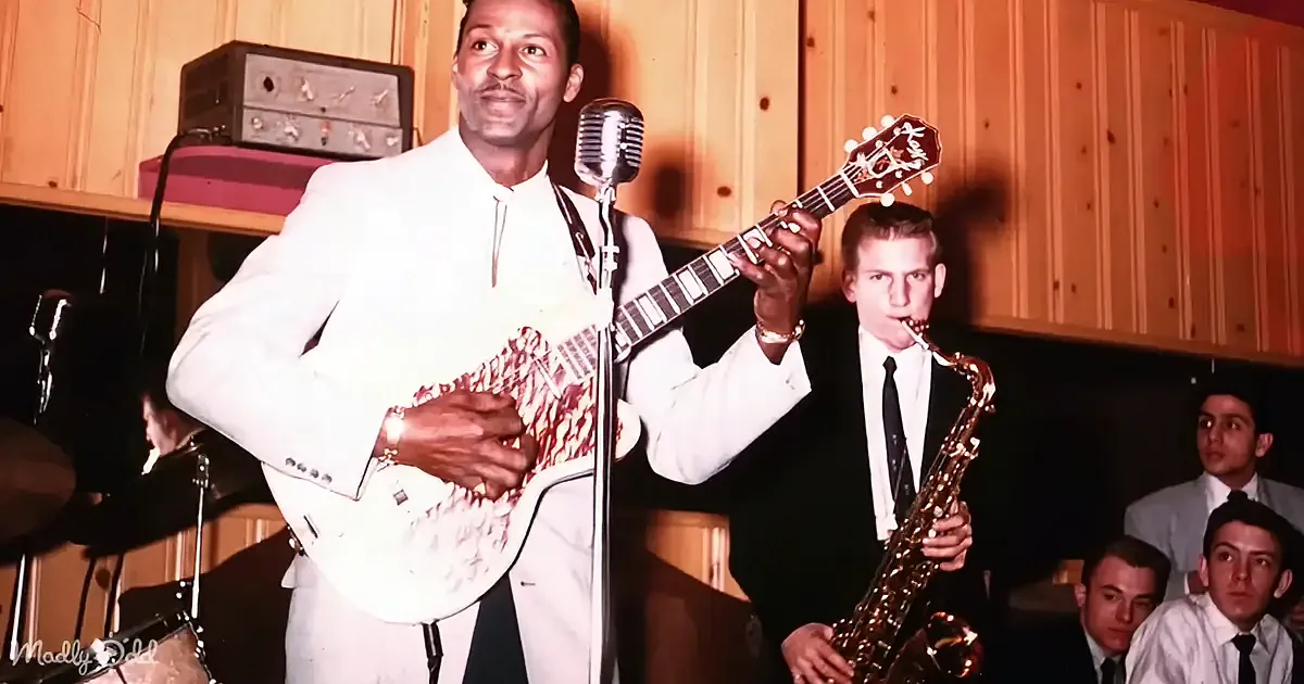 A man in a white suit plays a guitar on stage, surrounded by musicians and an audience.