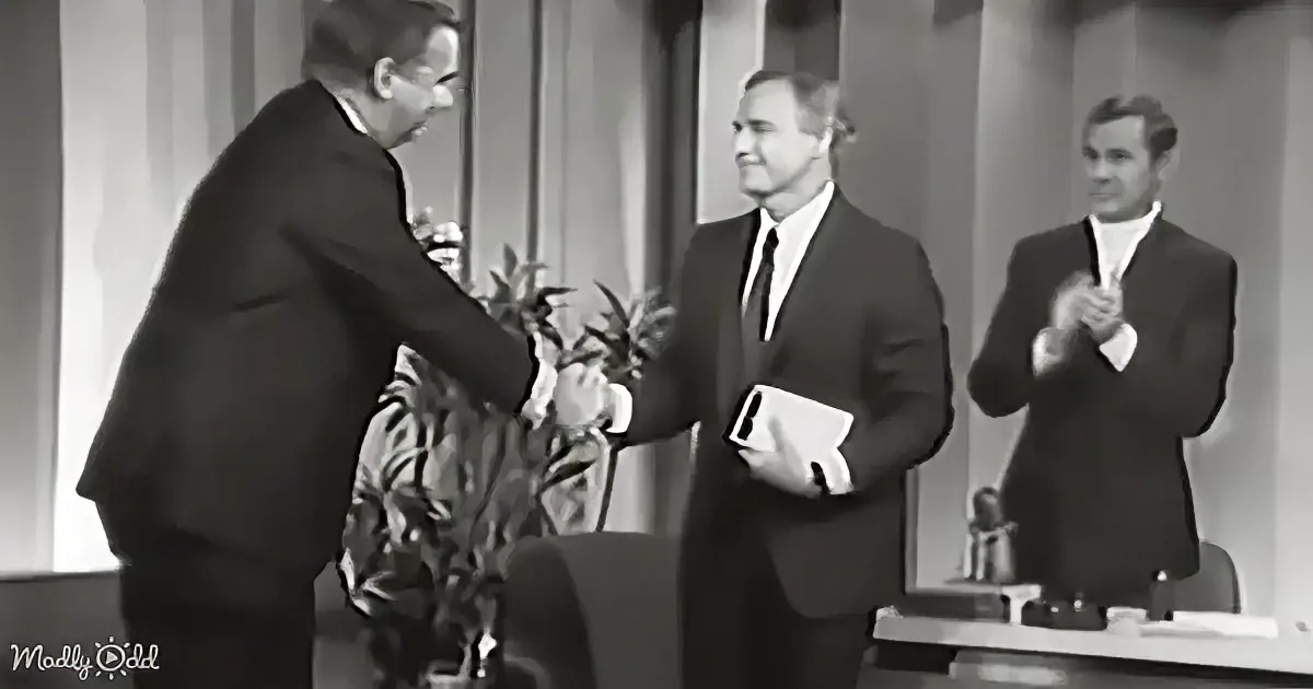 A man in a suit shakes hands with another man in a formal setting, while a third man claps in the background.