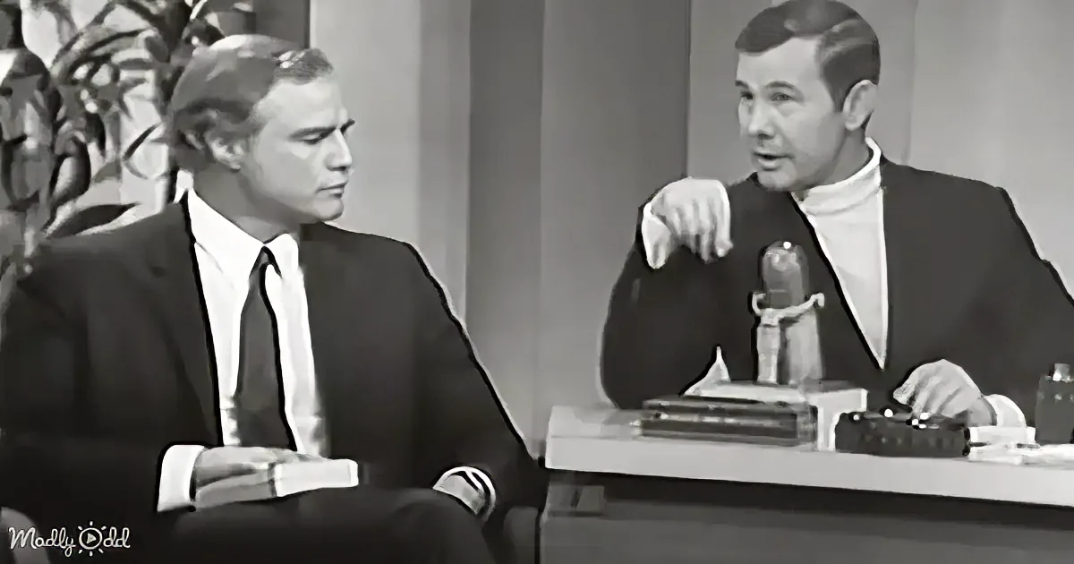 A man in a suit and tie sits at a desk, engaged in conversation with another man in a turtleneck.