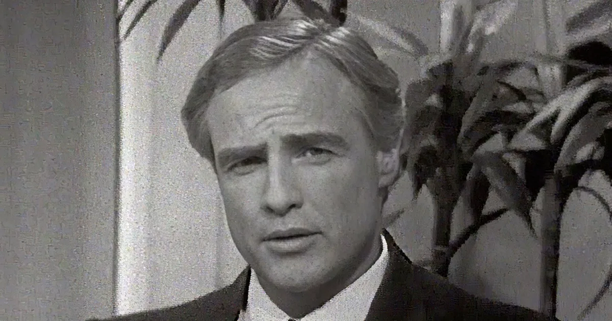 Two men in suits, one with a tie and the other in a turtleneck, both in black-and-white.