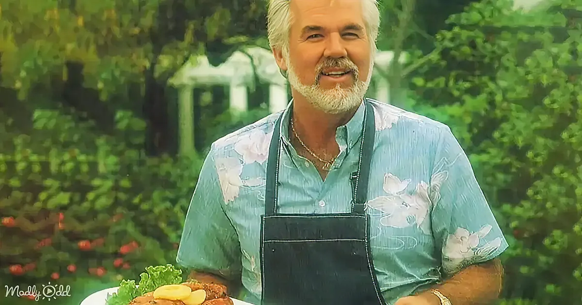 A man in a floral shirt and apron holds a plate of food, smiling in a garden setting.