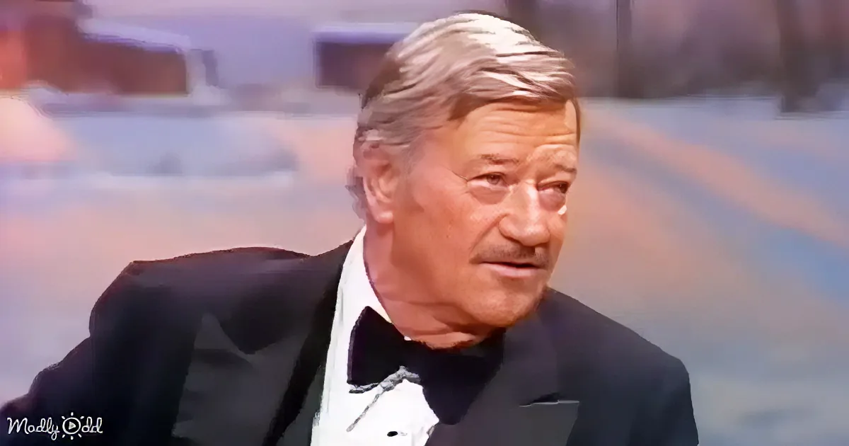 A man in a tuxedo with a bow tie is seated, looking to the side with a blurred background.