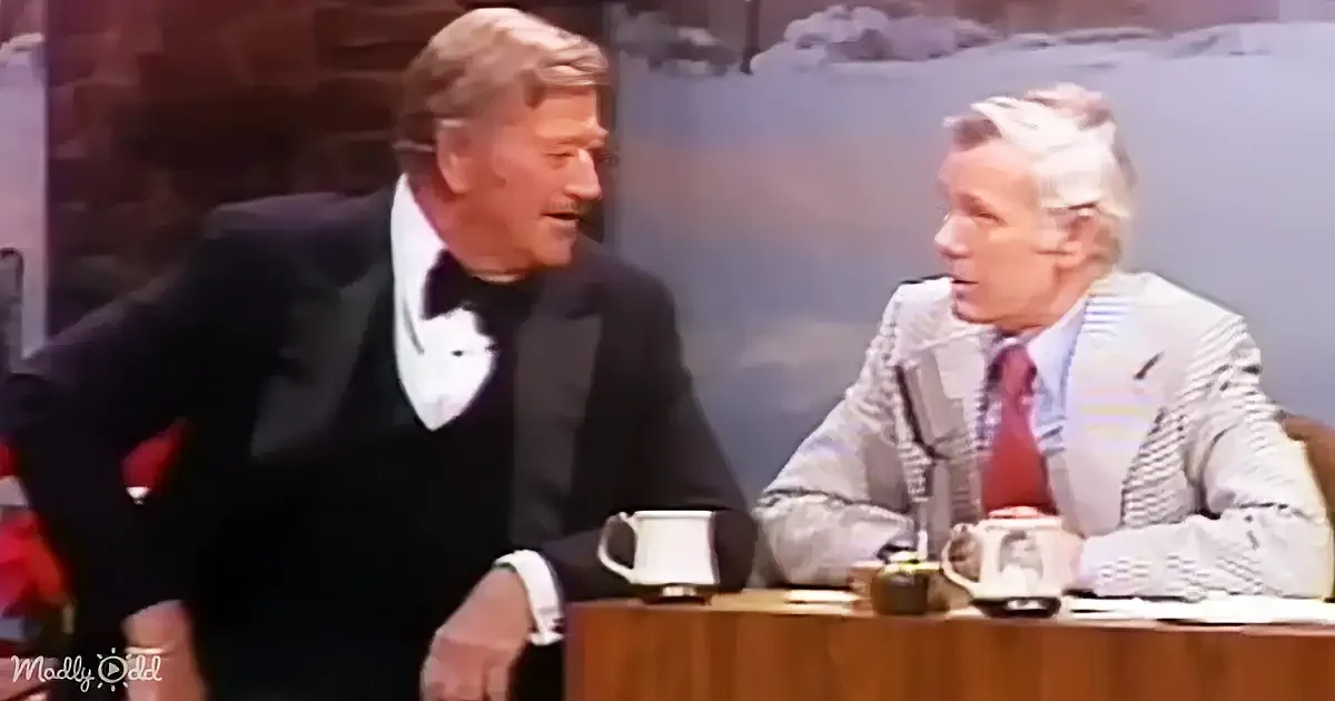 A man in a tuxedo leans toward another man in a striped suit at a desk with mugs.