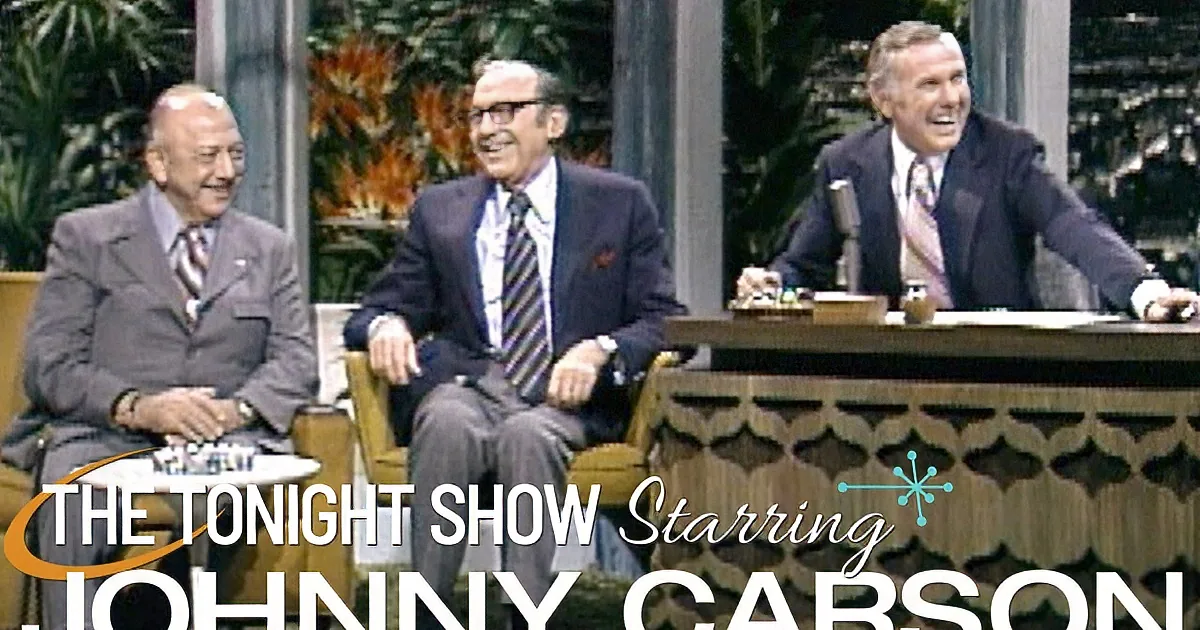Three men in suits are seated, one on a desk, discussing on a talk show set with tropical plants in the background.