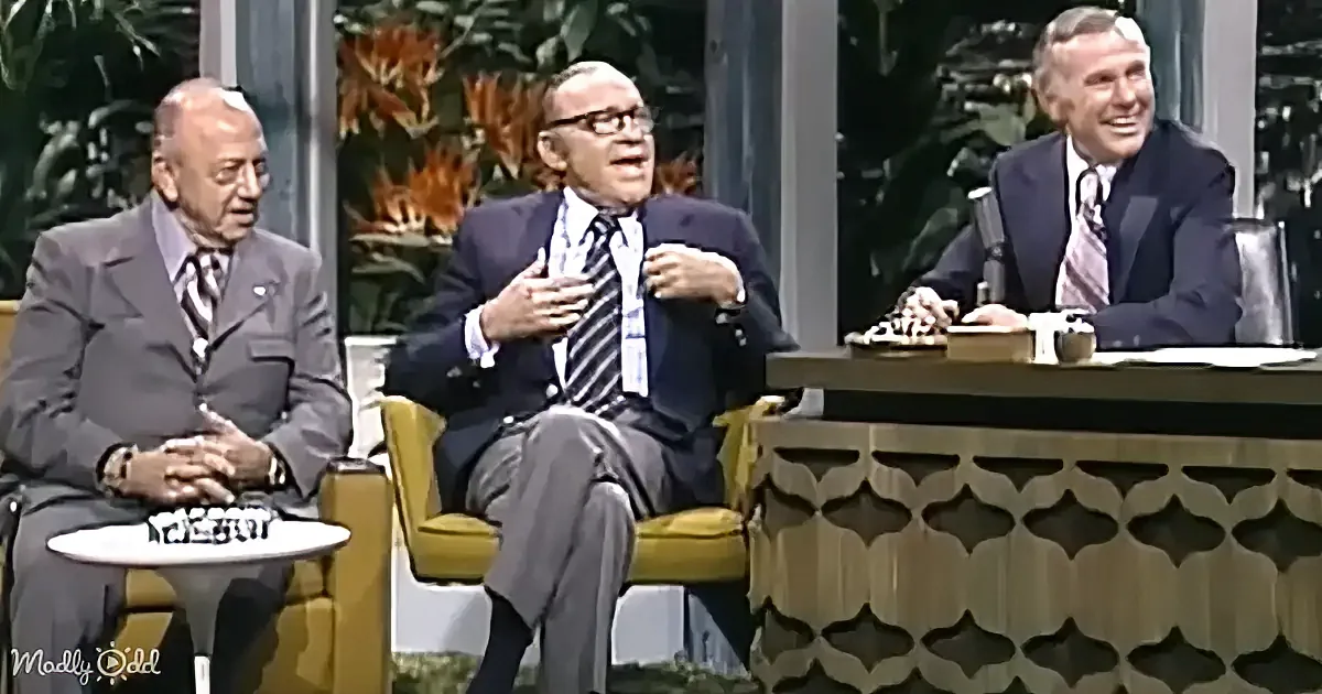 Three men in suits sit on a talk show set, one laughing, another gesturing, and the third watching.