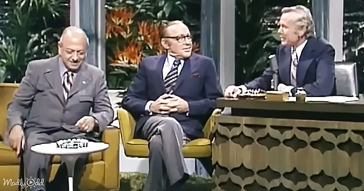 Three men in suits sit on a talk show set, one with a drink, another with hands clasped, and the third smiling.