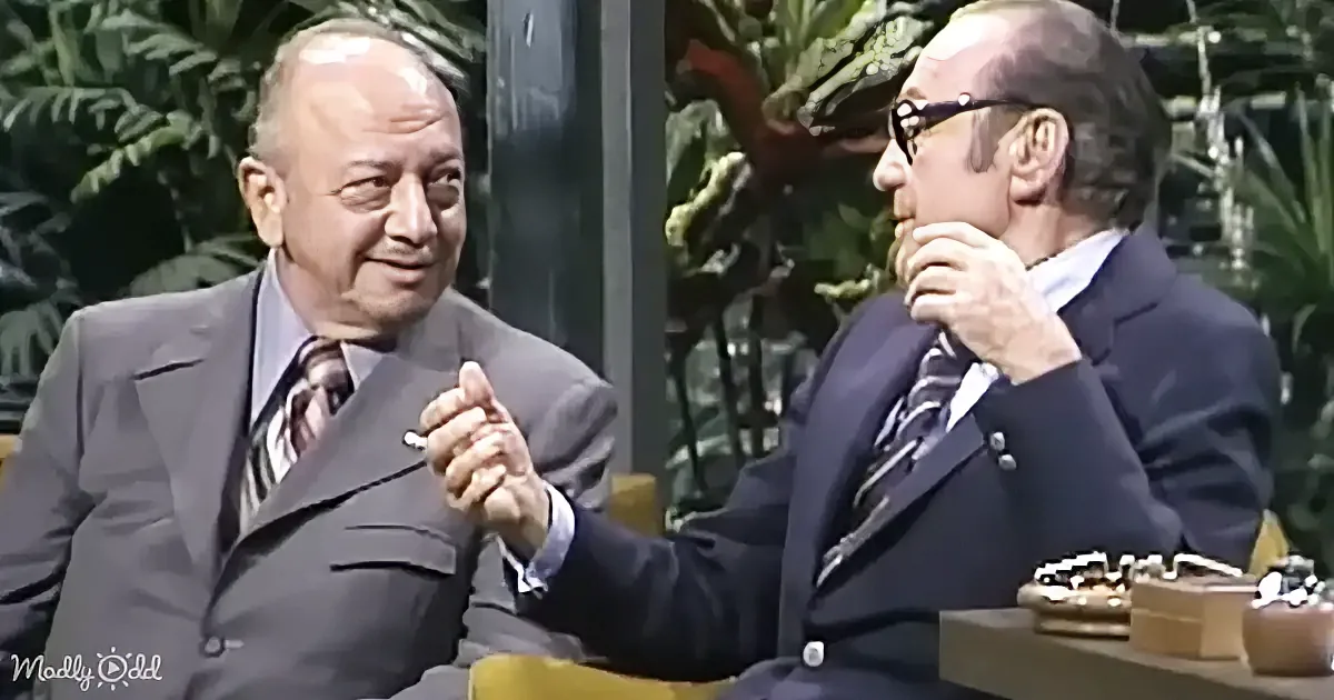 Two men in suits are seated, one gesturing while the other listens, in a setting with greenery in the background.
