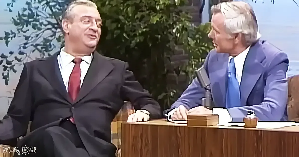 Two men in suits are seated at a desk, one in a red tie smiling, the other in a blue tie speaking.