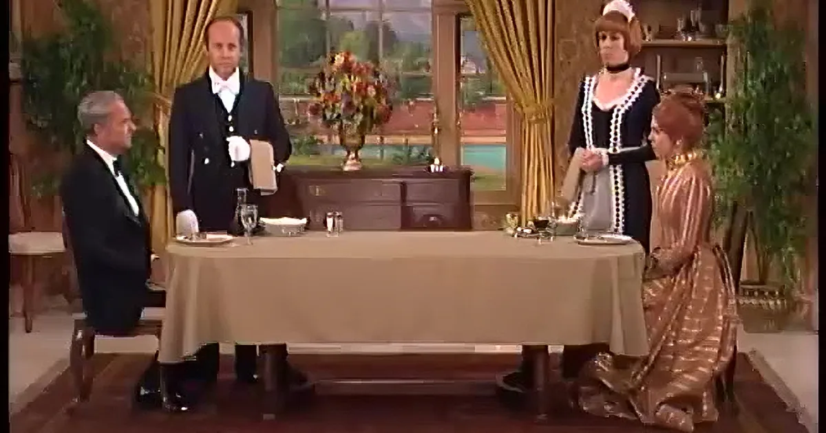 A man in a tuxedo stands at a dining table with a woman in a maid's uniform and another woman in a dress, all in a formal...