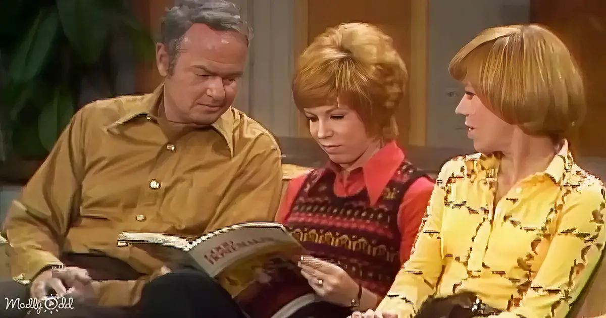 A man in a tan shirt reads a book while two women, one in a red and black sweater and the other in a yellow patterned...